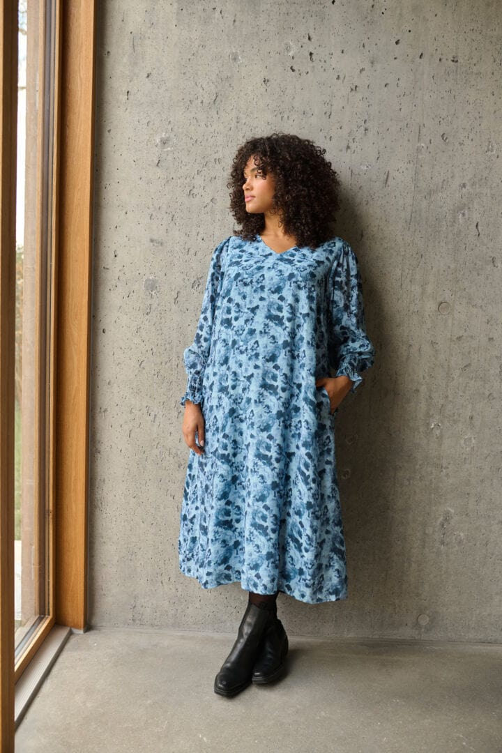 En frisk og herlig kjole i rett fasong med blått mønster og v-hals fra Kaffe Curve. Små rynker på skulderne som gir et løft og strikk i ermer så lengden kan justeres enkelt. En kjole som er klar for eventyr. 